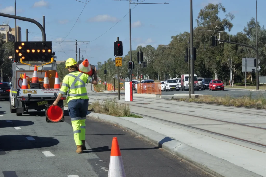 Traffic controller carrying cones beside a truck-mounted arrow board on an urban road with tram tracks