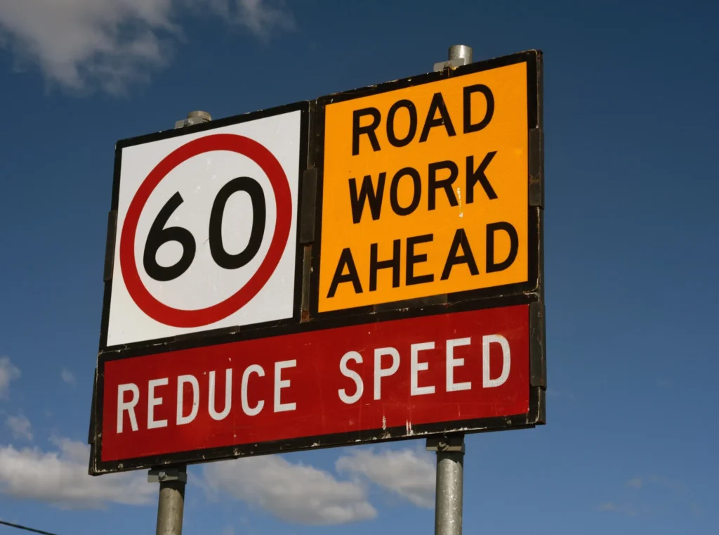 Roadside sign showing 60 km/h limit, ROAD WORK AHEAD and REDUCE SPEED against a blue sky