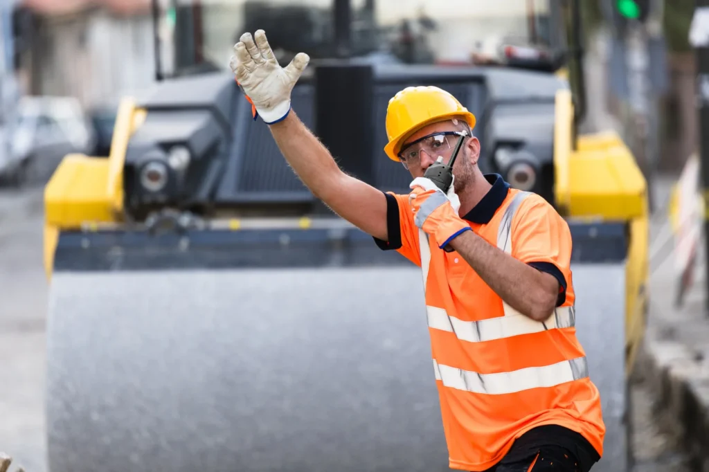 Road worker in hi-vis and hard hat signalling by hand in front of a road roller during paving