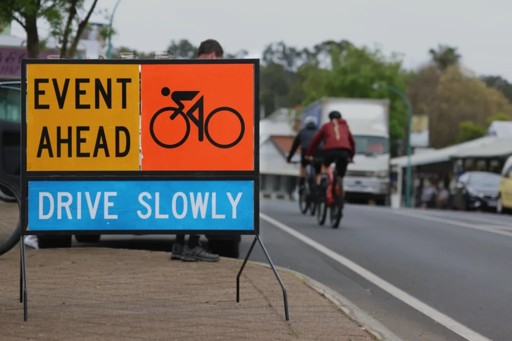 Roadside "Event ahead, drive slowly" sign with a cyclist symbol as cyclists ride past