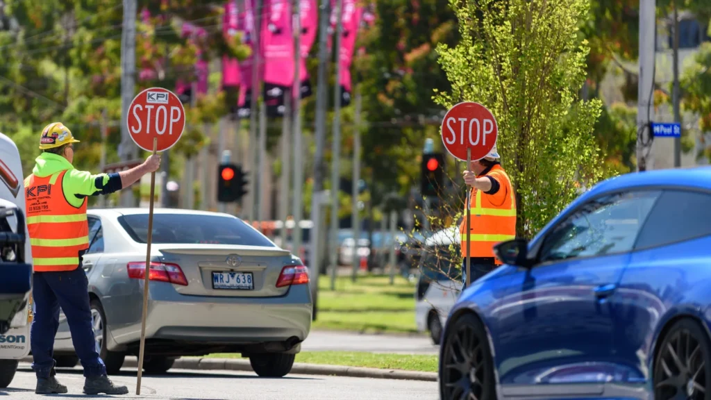 Two traffic controllers in hi-vis vests hold STOP signs at a road intersection