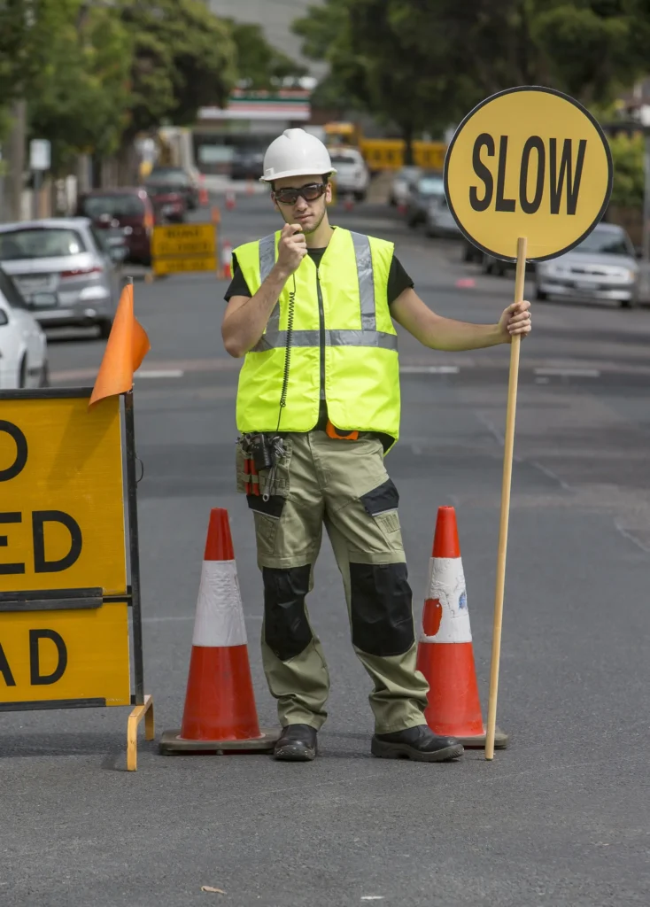 Traffic controller in hi-vis and hard hat holding a SLOW sign and blowing a whistle on a road